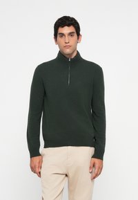 Uomo che indossa un maglione verde scuro con mezze zip e pantaloni beige, in piedi su uno sfondo bianco semplice, rivolto in avanti con un'espressione neutra.