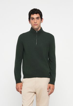 Hombre vestido con un suéter verde oscuro de media cremallera y pantalones beige, de pie contra un fondo blanco liso, mirando hacia adelante con una expresión neutral.