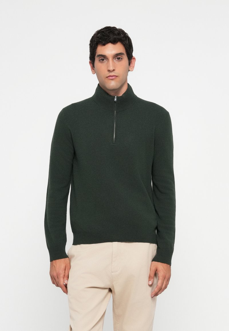 Uomo che indossa un maglione verde scuro con mezze zip e pantaloni beige, in piedi su uno sfondo bianco semplice, rivolto in avanti con un'espressione neutra.