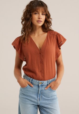 Ženska z valovitimi rjavimi lasmi, oblečena v rjavo-rdečo bluzo z gumbi in rokavi na nabirke ter svetlo modre kavbojke z visokim pasom, z rokami v žepih.