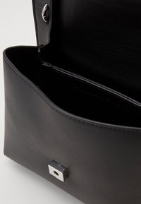 Černá kožená kabelka s otevřeným víkem, které odhaluje hladký interiér a čtvercovou stříbrnou sponu na přední straně.