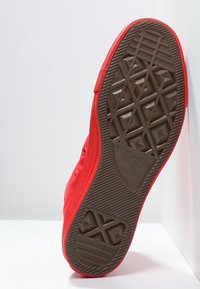 Sneaker di tela rossa con suola in gomma marrone texturizzata, caratterizzata da un motivo a diamante e punta liscia e arrotondata. Occhielli metallici sui lati.