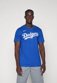 Maglietta in cotone blu con testo bianco "Dodgers", maniche corte e logo Nike sulla spalla sinistra. Indossata con pantaloni corti grigio chiaro.