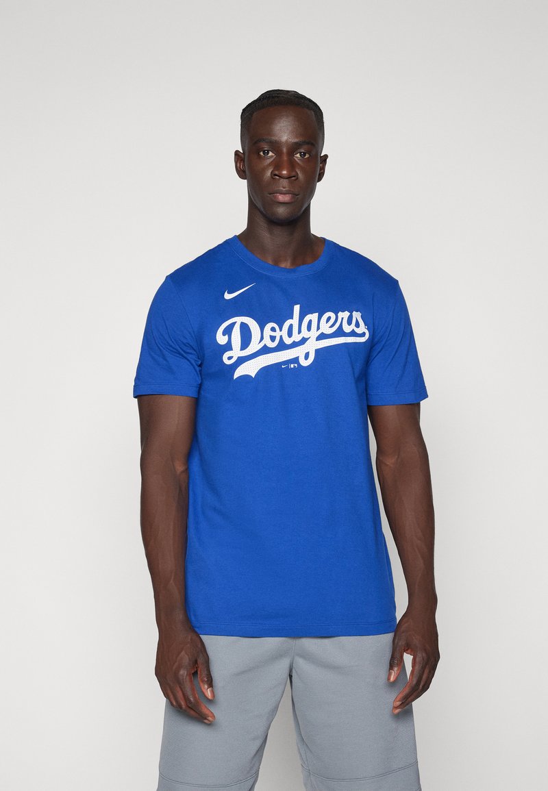 Maglietta in cotone blu con testo bianco "Dodgers", maniche corte e logo Nike sulla spalla sinistra. Indossata con pantaloni corti grigio chiaro.
