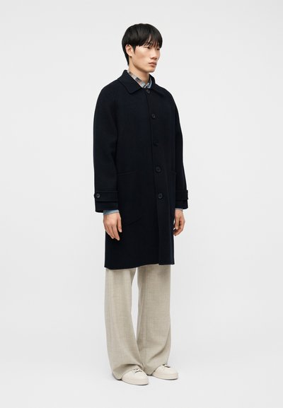 Άνδρας που φοράει μαύρο παλτό με κουμπιά  πάνω από πουκάμισο με καρό σχέδιο, μπεζ παντελόνι με φαρδιά πόδια και λευκά αθλητικά παπούτσια, στέκεται μπροστά από απλό φόντο.