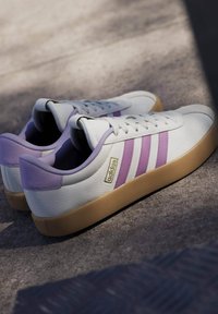 Zapatillas de cuero blanco con detalles morados, tres franjas y suela de goma; cuenta con un acabado texturizado y un cuello acolchado.