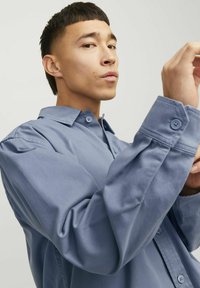 Chemise bleu clair à manches longues avec un col boutonné. Elle possède une poche avant et des boutons aux poignets. Le tissu semble lisse avec un léger éclat.