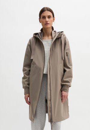 Kobieta ubrana w długi taupe płaszcz z kapturem i zamkiem z przodu, na jasnoszarej bluzie i spodniach dresowych, stojąca na jednolitym tle.