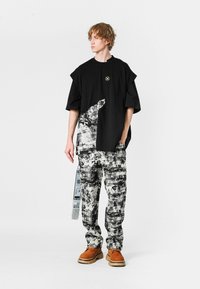 Joven vestido con una camiseta negra de gran tamaño con gráfico, pantalones abstractos en blanco y negro, y zapatos marrones de suela gruesa, de pie frente a un fondo blanco liso.