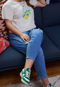 Personne assise sur un canapé bleu portant un t-shirt beige avec un imprimé citron, un jean bleu déchiré et des baskets vertes et blanches.