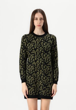 Femme portant une robe noire à manches longues avec un imprimé animal abstrait vert, debout devant un fond blanc.