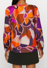 Blusa multicolorida con grandes patrones florales en naranja, púrpura y marrón, con mangas largas y una costura fruncida en la parte posterior.