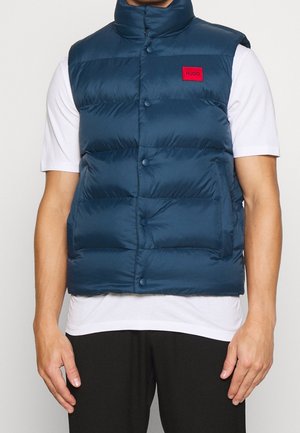 Mann trägt eine marineblaue gesteppte, ärmellose Steppweste über einem weißen kurzärmligen Hemd, mit einem roten rechteckigen Logoaufnäher auf der Brust.