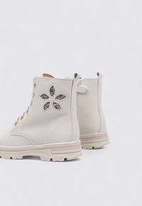 Une paire de bottines en cuir blanc cassé avec des découpes en forme de fleurs remplies de paillettes et des semelles en caoutchouc chunky beiges.