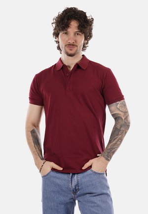 Uomo con capelli ricci che indossa una polo bordeaux e jeans blu, mani in tasca, che mostra tatuaggi su entrambe le braccia su uno sfondo uniforme.