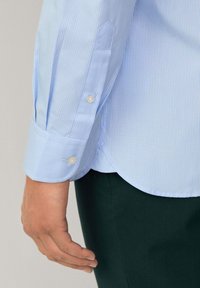 Light blue striped long-sleeve shirt with buttoned cuffs, featuring white buttons and a rounded hem. Fabric appears smooth and soft.