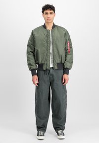 Maslinasto zelena bomber jakna s prednjim zatvaračem i rebrastim manžetama, uparena s prostranim tamnosivim cargo hlačama i crno-bijelim tenisicama.