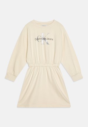 Robe pull crème avec taille élastique, manches longues et impression de logo argenté sur la poitrine. Texture de tissu douce et lisse.