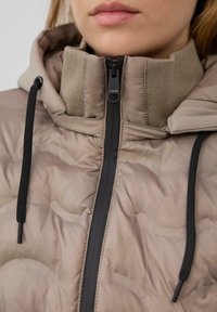 Chaqueta acolchada de color marrón claro con un cuello alto, que presenta una cremallera negra, cordones y acentos de tela texturizada.