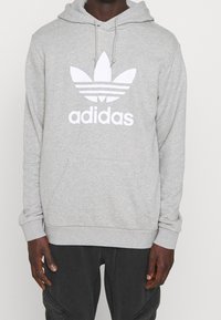 Grå hoodie med en framficka, som har en stor vit Adidas trefoil-logga. Tillverkad av mjukt, texturerat material med justerbara snören.