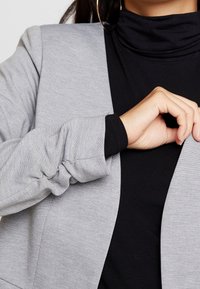 Harmaa bleiseri, jossa on ribbikuvio, pitkät hihat ja istuva malli. Päällä musta poolopaita.