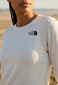 Donna con capelli intrecciati che indossa una maglietta bianca a maniche lunghe con il logo The North Face su sfondo all'aperto.