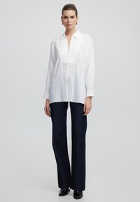 Camicia bianca a maniche lunghe con scollo a V e chiusura con bottoni; abbinata a jeans svasati blu scuro; tessuto liscio e leggero.