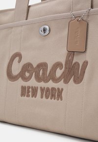 Sac cabas en toile beige avec "Coach New York" brodé en marron et une étiquette en cuir assortie attachée par une chaîne à billes argentée.