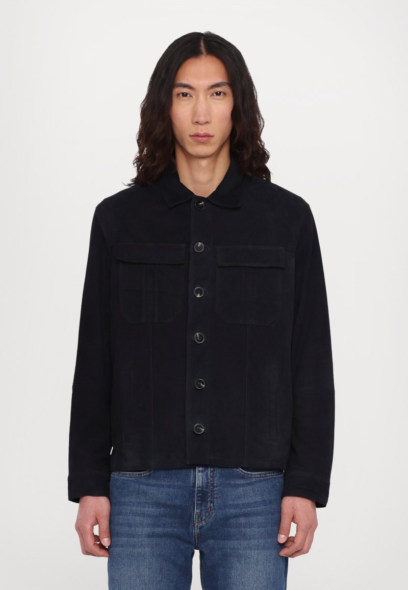 Veste noire en tissu lisse avec un devant à boutons, deux poches poitrine et un col. Portée avec un jean en denim bleu.