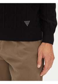 Schwarzer, strukturierter Pullover mit gerippten Bündchen und Saum, versehen mit einem grauen, dreieckigen Logopatch. Kombiniert mit sichtbarer beiger Hose im unteren Bereich.
