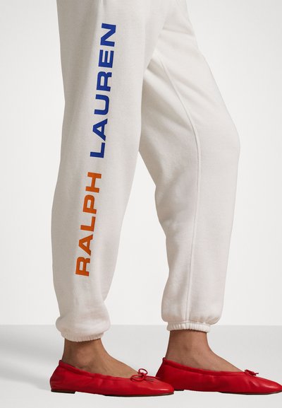 Λευκά φόρμας με μπλε και πορτοκαλί κείμενο "RALPH LAUREN" στο πλάι, συνδυασμένα με κόκκινες φλατ παπούτσια με λεπτομέρεια φιόγκου. Ομαλή επιφάνεια φόντου.