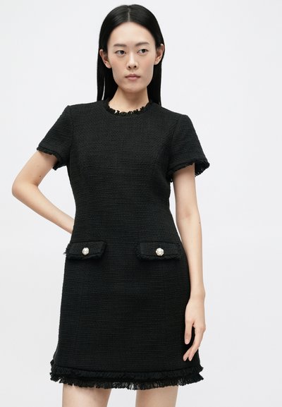 Rochie neagră texturată cu mâneci scurte, margini desfăcute, două buzunare frontale cu butoane din perlă și decolteu rotund.
