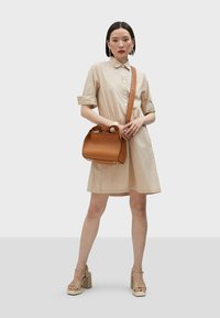 Abito camicia color beige con maniche corte, chiusura a bottoni, abbinato a una borsa a tracolla in pelle marrone con parte superiore intrecciata. Sandali con piattaforma verdi.