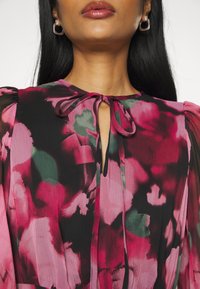 Femme portant un chemisier floral transparent avec des motifs abstraits rose, rouge et vert, doté d'un col à nœud et de manches bouffantes.