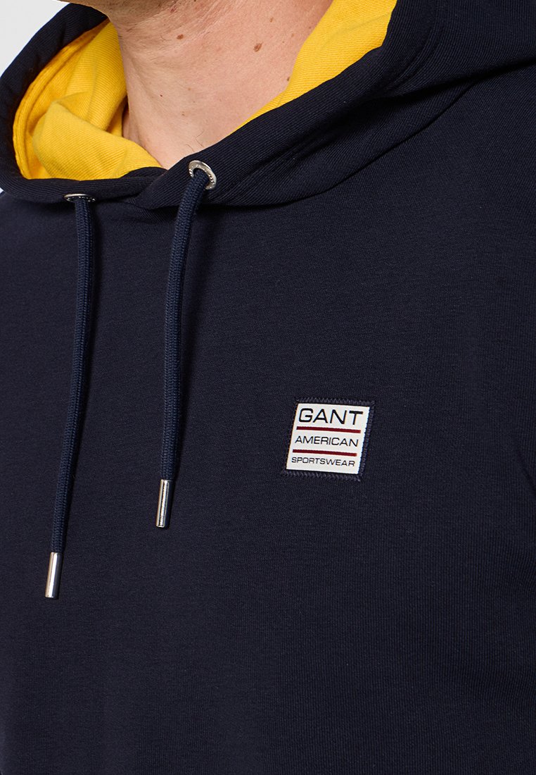 Sweat à capuche bleu marine avec une doublure intérieure jaune, comportant un patch logo sur la poitrine et des éléments en métal argenté pour les cordons.