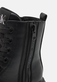 Svart lærstøvlett med snøring foran og glidelås på siden. Har teksturert materiale og "Calvin Klein Jeans"-merking i sølv.