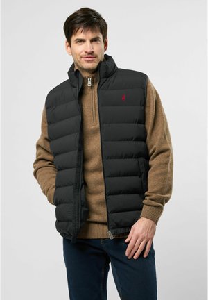 Melnā piepūšamā veste ar augstu apkakli un priekšējo rāvējslēdzēju, uzvilkta virs brūnas adītas džemperis. Ar mazu sarkanu logotipu uz krūtīm.