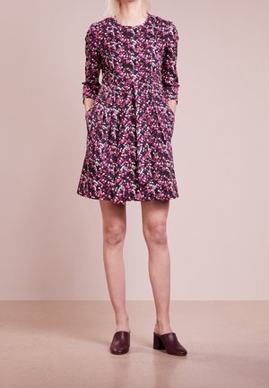 Frau trägt ein knielanges, langärmliges Blumenkleid mit pinken, weißen und violetten Blumen, kombiniert mit dunkelvioletten Mules mit Absatz.