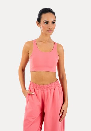 Frau mit dunklen Haaren im Dutt, trägt ein pinkes ärmelloses Crop-Top und passende lockere Hose, steht mit einer Hand in der Tasche vor weißem Hintergrund.