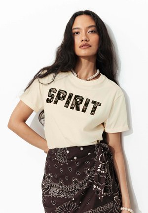 Femme aux longs cheveux foncés portant un t-shirt beige "SPIRIT", un collier de perles et une jupe portefeuille noire à motifs sur fond blanc.