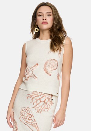 Frau trägt ärmelloses cremefarbenes Strickoberteil und Rock mit Korallen- und Muschelmuster sowie große goldene Scheibenohrringe.