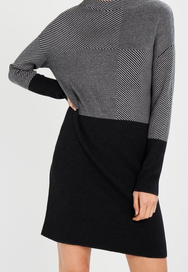 Femme portant une robe en tricot à manches longues, longueur genou, avec des sections géométriques en noir, gris foncé et gris clair, ainsi que des motifs rayés.