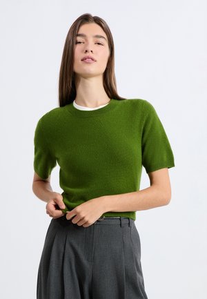 Grüner Kurzarmpullover aus weichem Material mit Rundhalsausschnitt. Kombiniert mit hoch taillierter grauer Hose mit Falten und Gürtel.