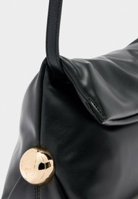 Zwarte leren handtas met een zachte, slungelige vorm en een ronde gouden metalen logo-versiering nabij de basis van de handgreep.