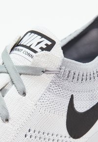 Buty Nike z białą i jasnoszarym dzianinowym cholewką, czarnym logo w kształcie smoka i szarymi sznurowadłami. Teksturowana powierzchnia z wyściełanym kołnierzem.
