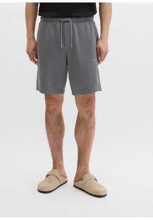 Mężczyzna w szarych szortach z sznurkiem i beżowych butach wsuwanych, stojący na tle jednolitego białego tła.