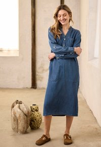 Une femme se tient les bras croisés, vêtue d'une robe en jean bleu et de chaussures marron, dans une pièce minimaliste avec deux vases en céramique texturée posés sur le sol.