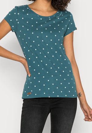 Frau trägt ein türkisfarbenes kurzärmeliges Shirt mit weißen Punkten und schwarze Jeans, Hand auf der Hüfte, kleines Tattoo am rechten Handgelenk.