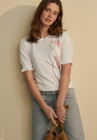 Witte kortaf mouwtrui met een bloemenpatroon in roze en groen. Ribgebreide zoom en manchetten. Gecombineerd met lichtblauwe denim jeans en een bruine tas.