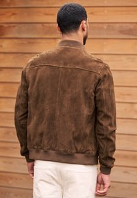 Mann mit kurzen Haaren, der eine braune Wildlederjacke und weiße Hose trägt und mit dem Rücken zur Kamera vor einer Holzwand steht.